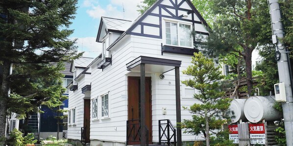 A white building with a dark roof and a small porch. There are trees and a utility pole with pipes visible.