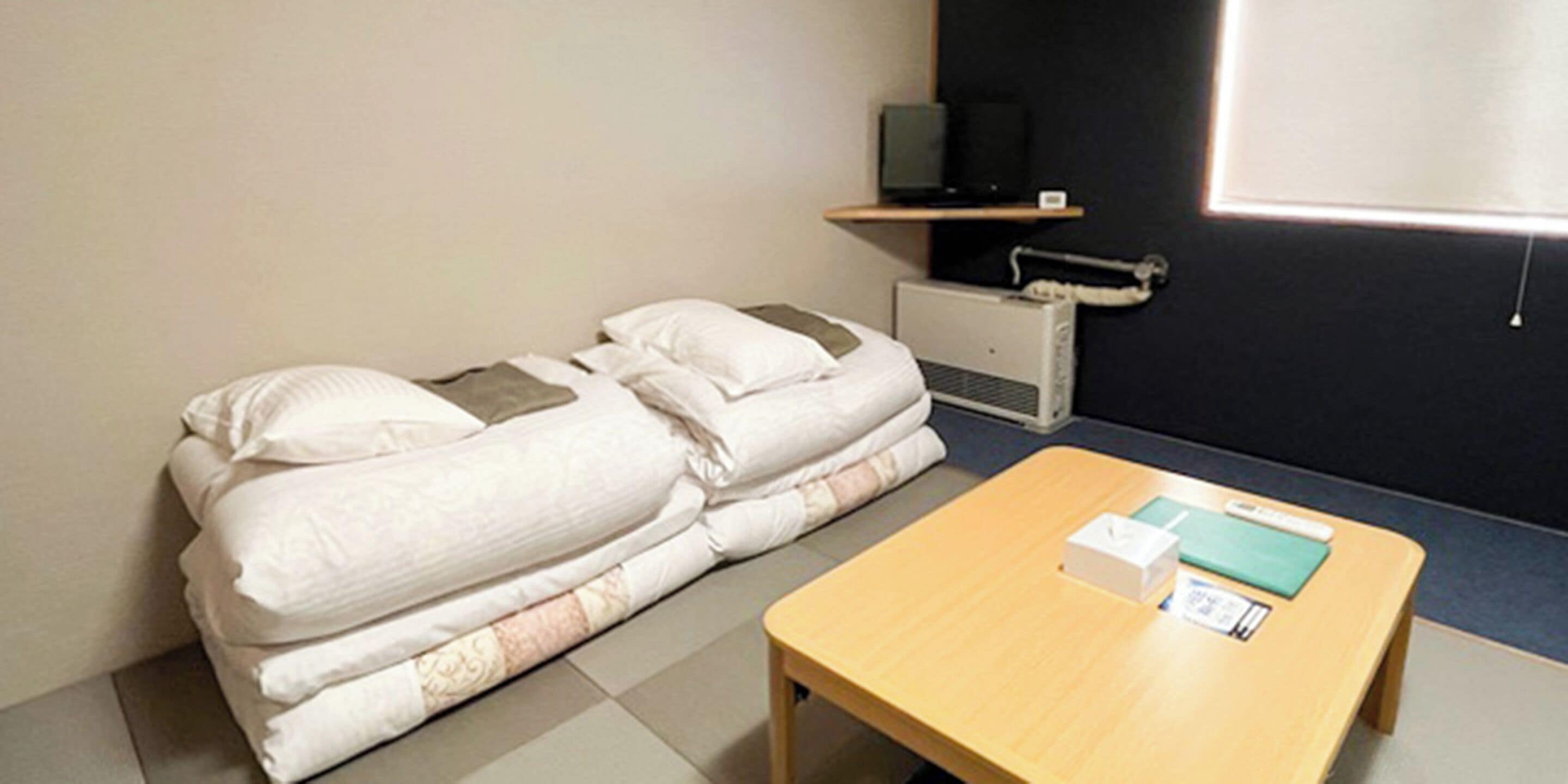 Hotellihuone, jossa on sänky, pieni pöytä, hyllyllä oleva televisio ja lämmitin.