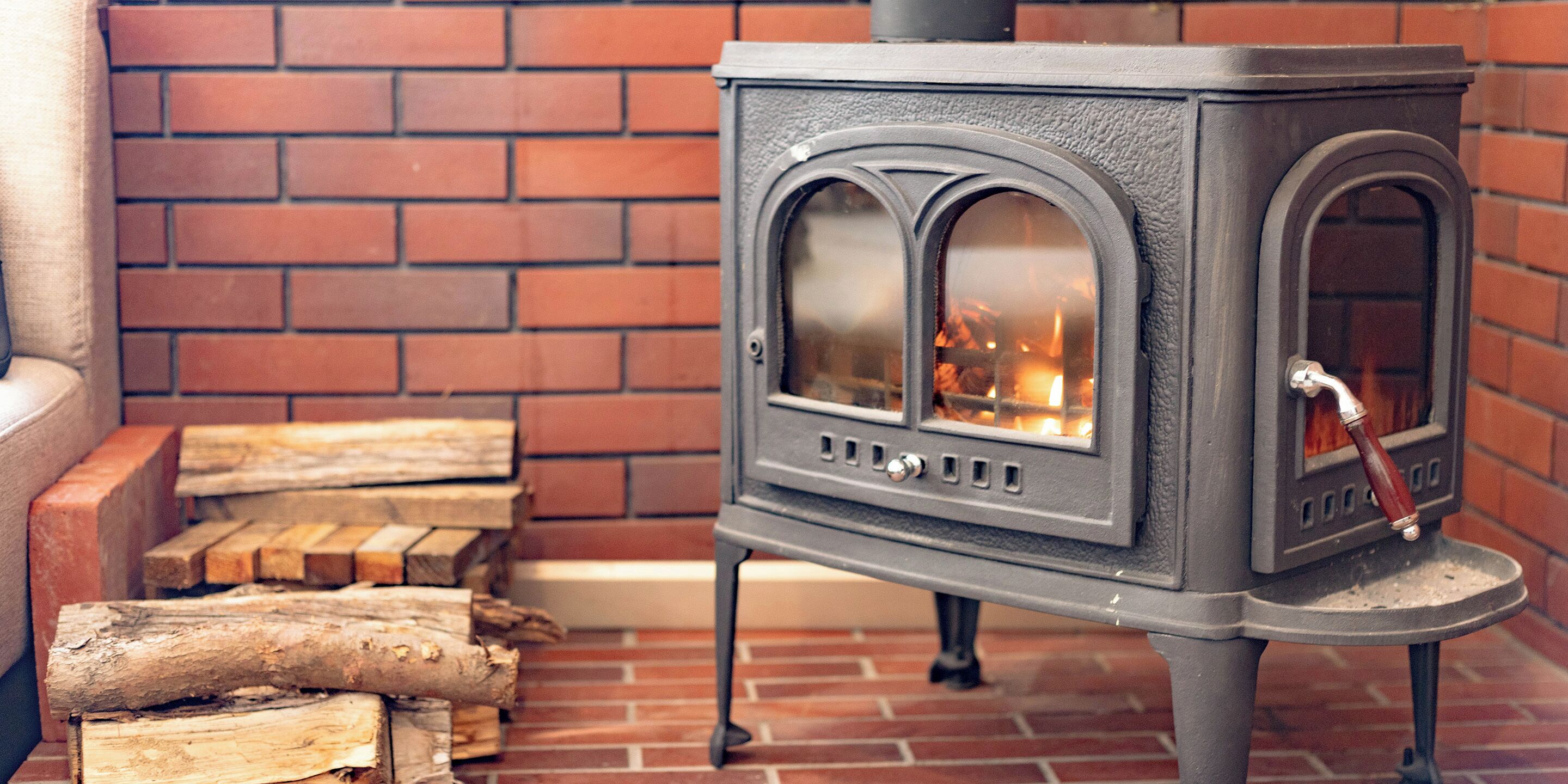 En vedeldad kamin med en glaslucka, placerad mot en tegelvägg med staplade vedträn i närheten.