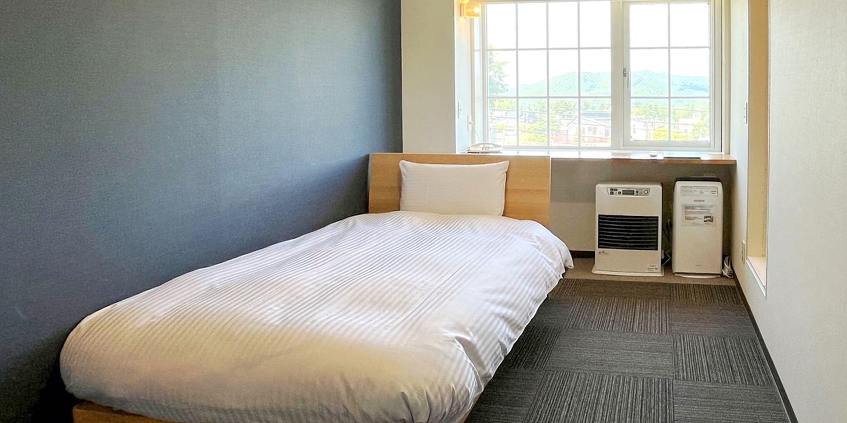 Una cama individual con almohada blanca, una ventana con vistas y dos equipos de aire acondicionado.