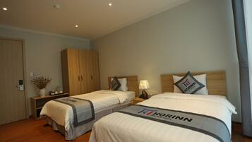 A hotel room with two beds, wooden wardrobe, bedside tables with lamps, and a vase with flowers on a shelf.