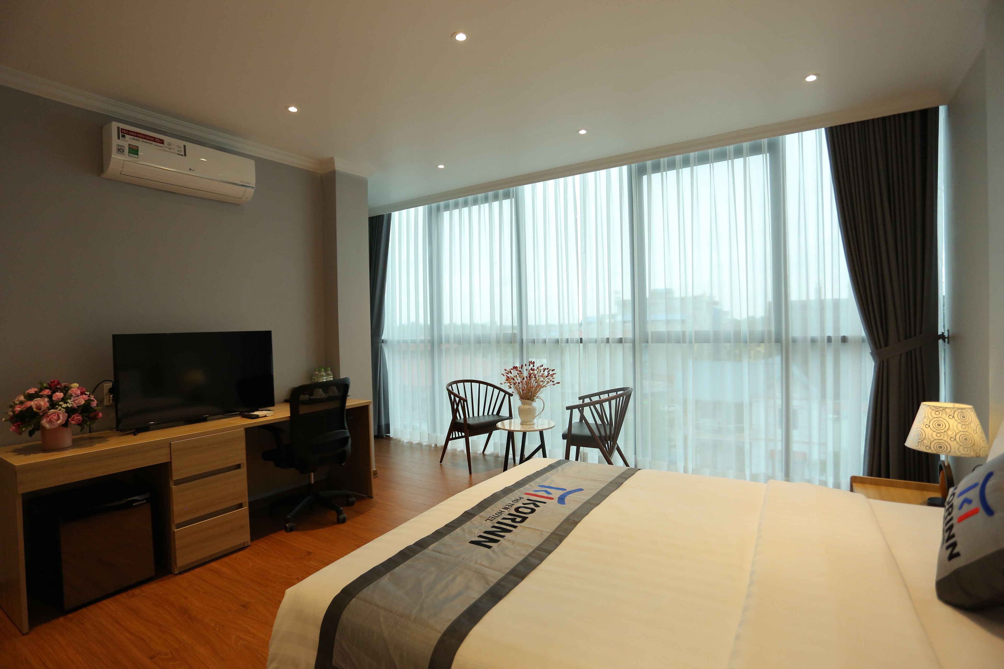 A hotel room with a bed, a desk with a TV, a chair, a small table with flowers, and a window with curtains.
