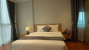 A hotel room with a large bed, a nightstand with a lamp, a pillow with the hotel's logo, and a flower vase.