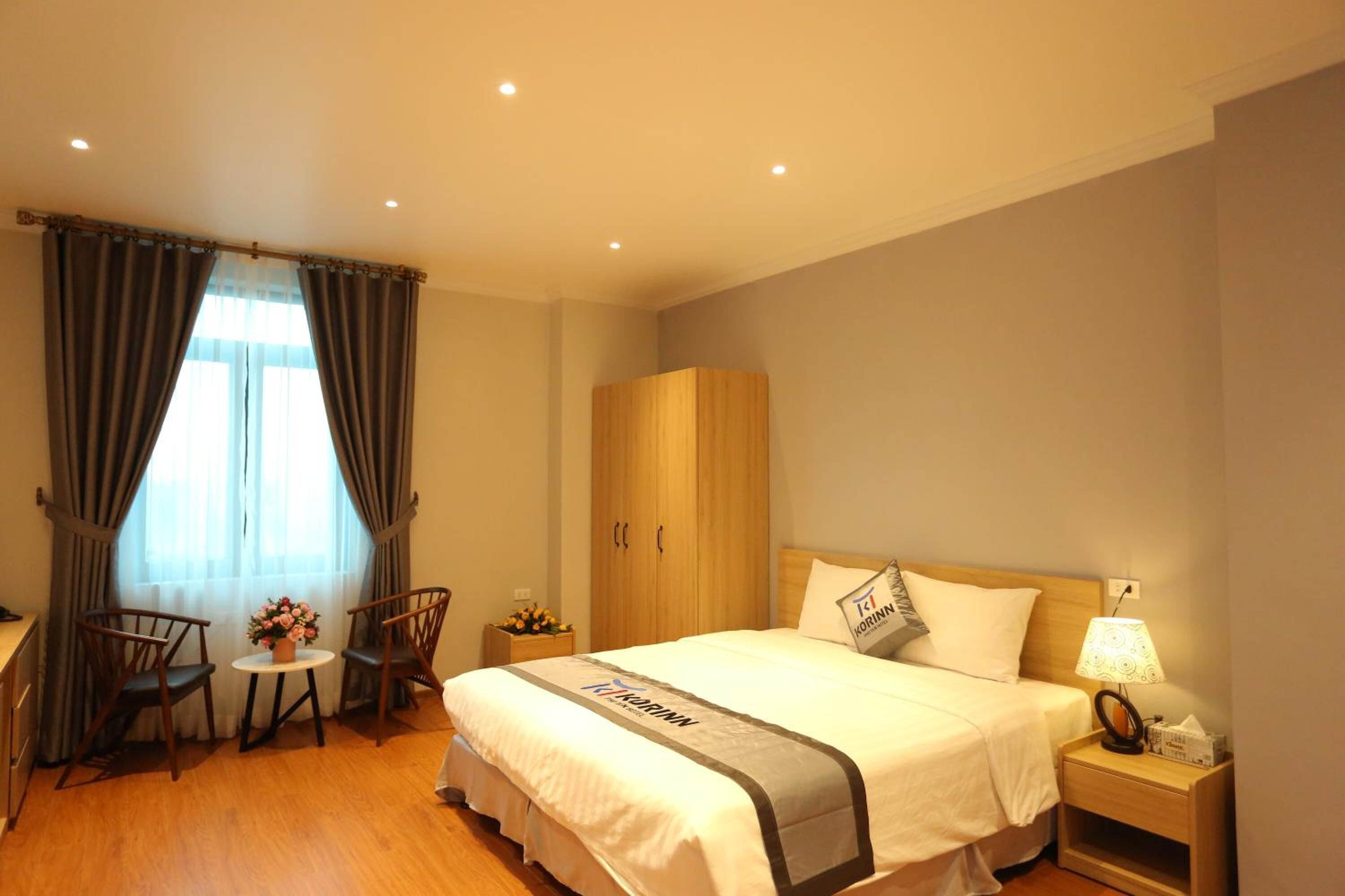 Een hotelkamer met een bed, een houten kledingkast, een bureau met stoel, een klein tafeltje met bloemen en een nachtkastje met lamp.
