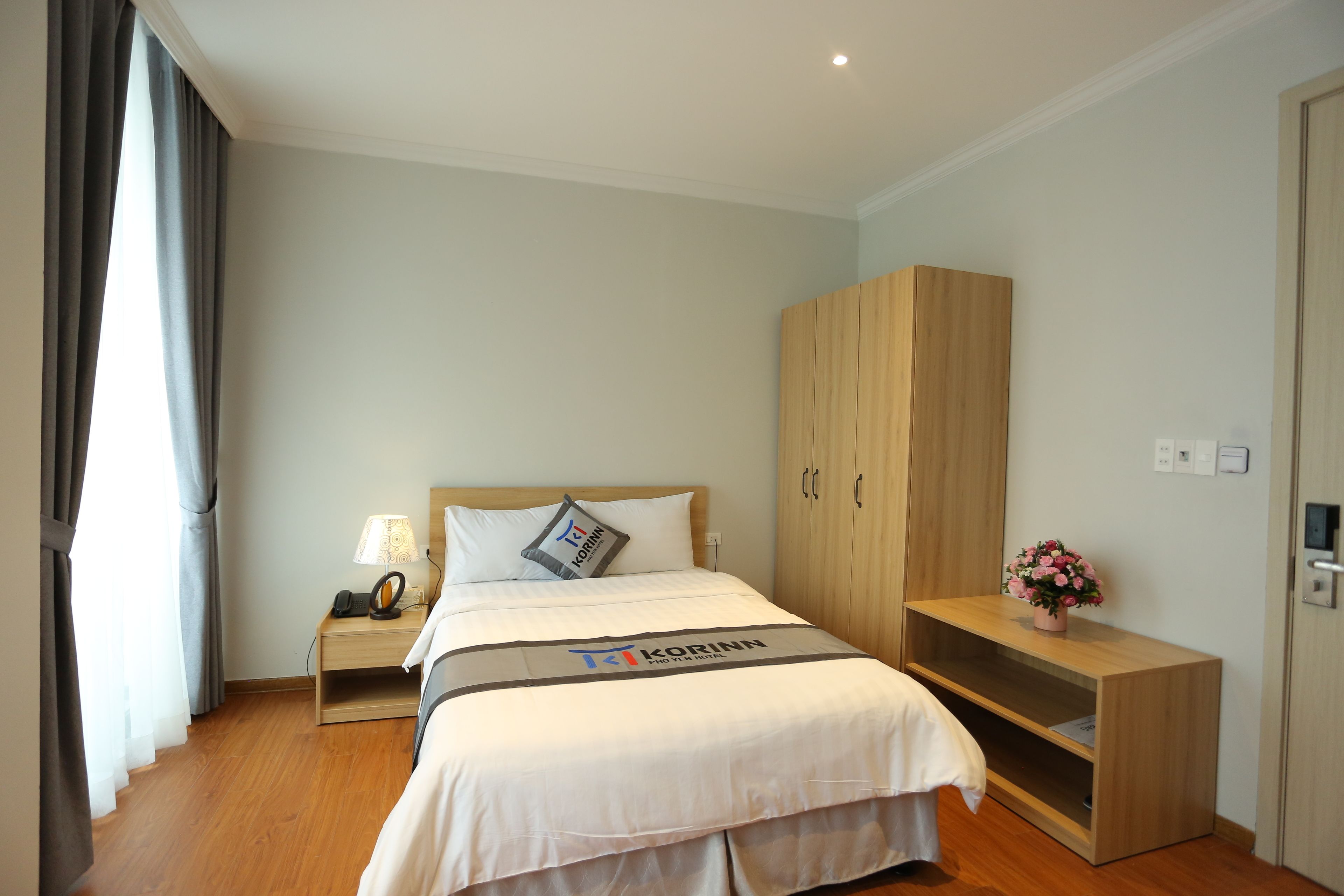 A hotel room with a bed, a nightstand, a wooden wardrobe, and a small table with a lamp and a flower vase.