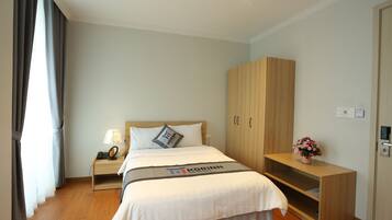 A hotel room with a bed, a nightstand, a wooden wardrobe, and a small table with a lamp and a flower vase.
