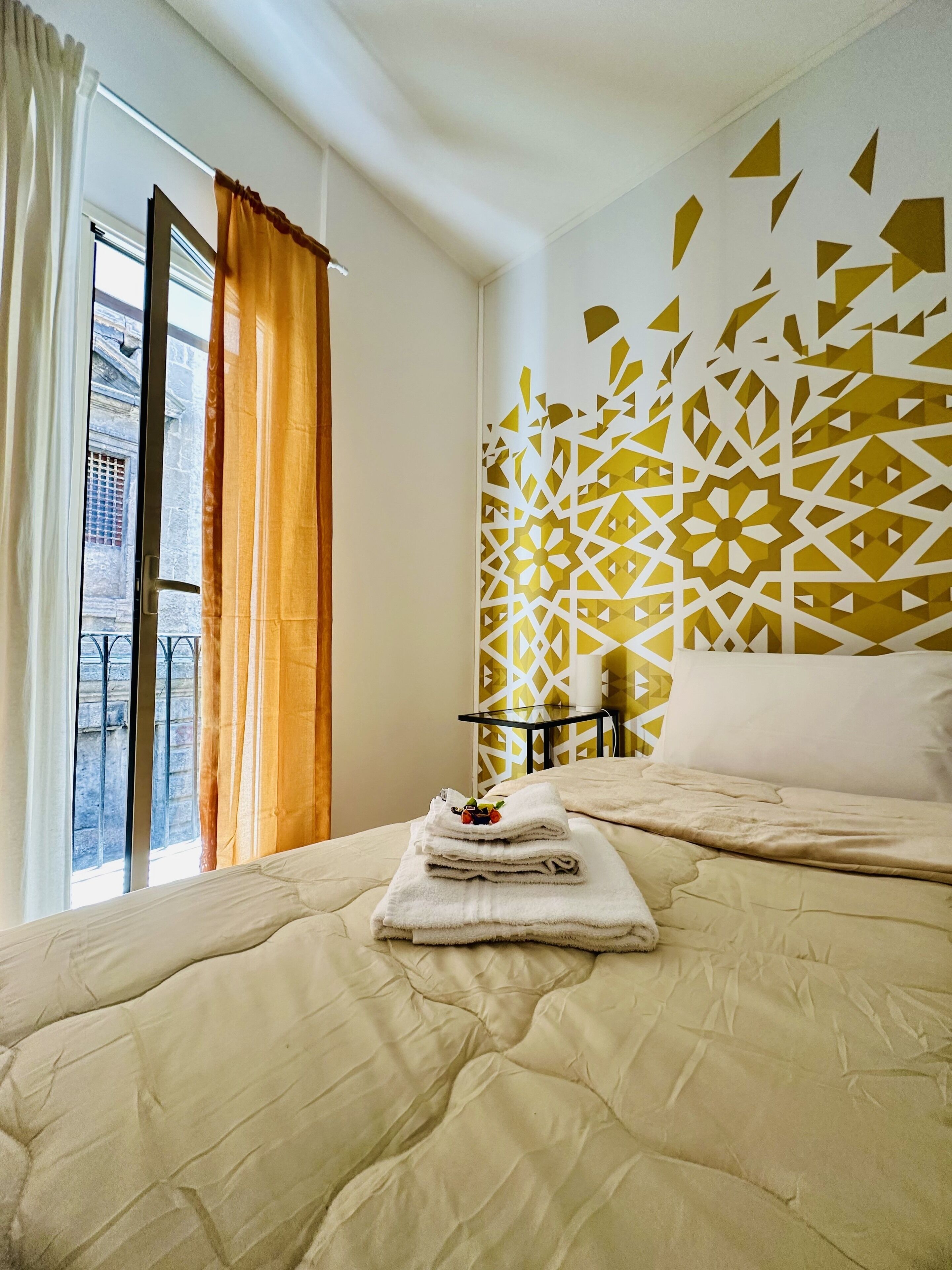 Une chambre d’hôtel avec un lit, une fenêtre avec des rideaux, un mur décoratif et une petite table avec une lampe.