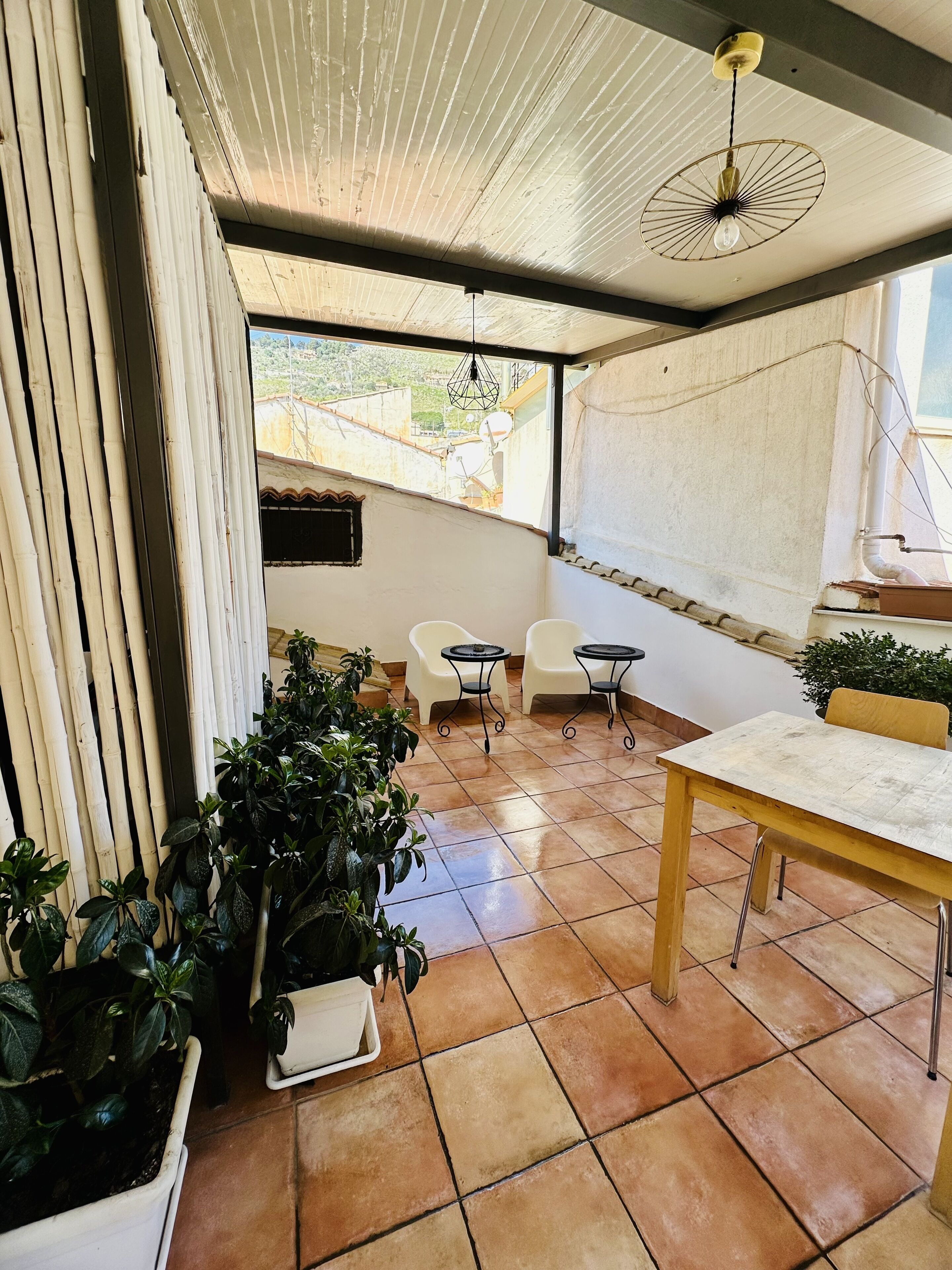 Ein Patio mit Fliesenboden, einem Holztisch, zwei weißen Stühlen und Topfpflanzen.