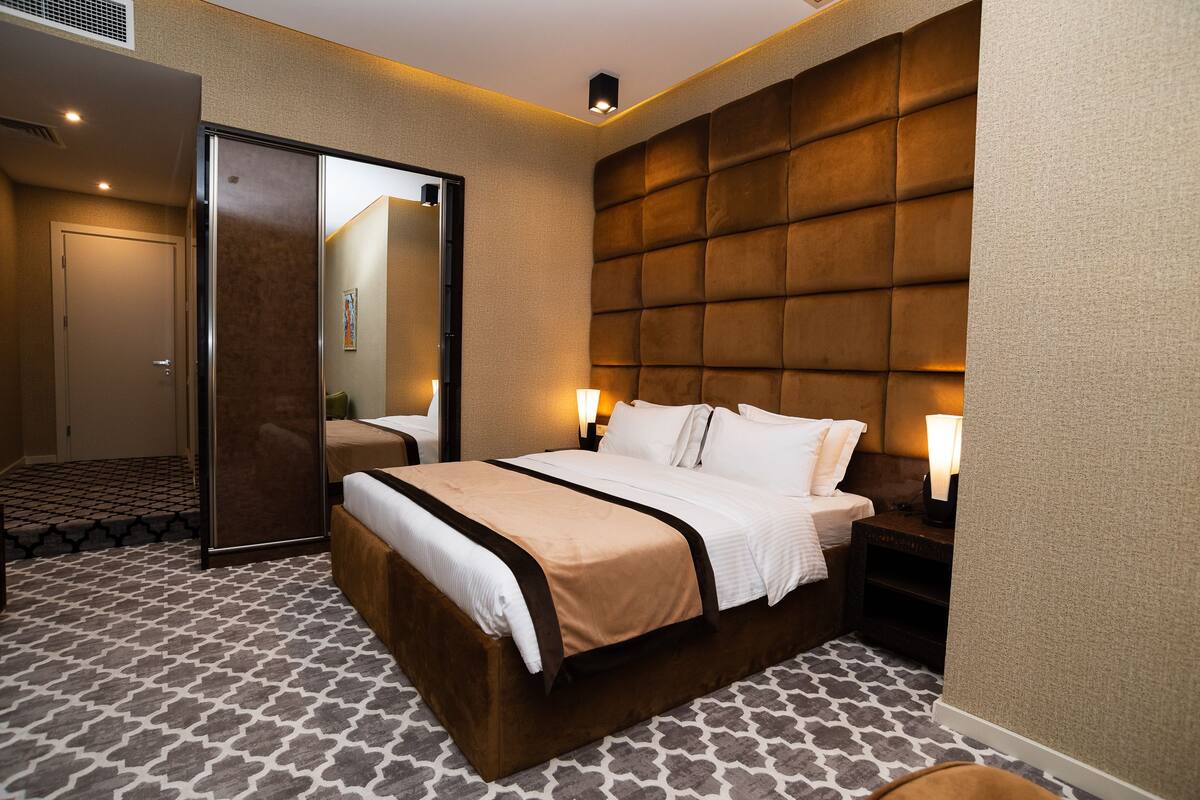 A hotel room with a large bed, a wardrobe, a bedside table with a lamp, and a patterned carpet.