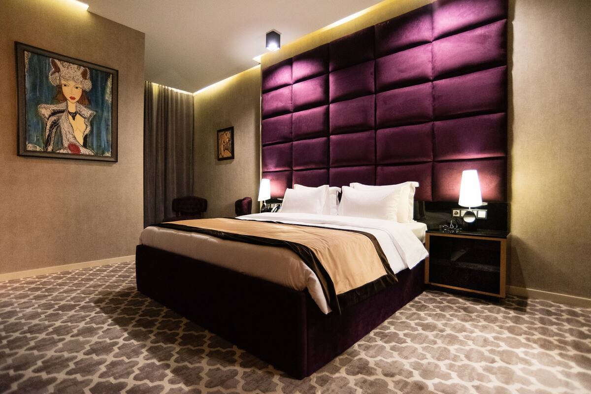 A hotel room with a large bed, a headboard, a nightstand, a lamp, and a painting on the wall.