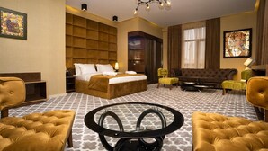 Quarto de hotel com uma cama grande, um sofá, duas poltronas, uma mesa de centro redonda e um lustre.