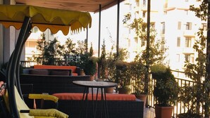 Varanda com móveis de vime, plantas em vasos e vista para um edifício ao fundo.