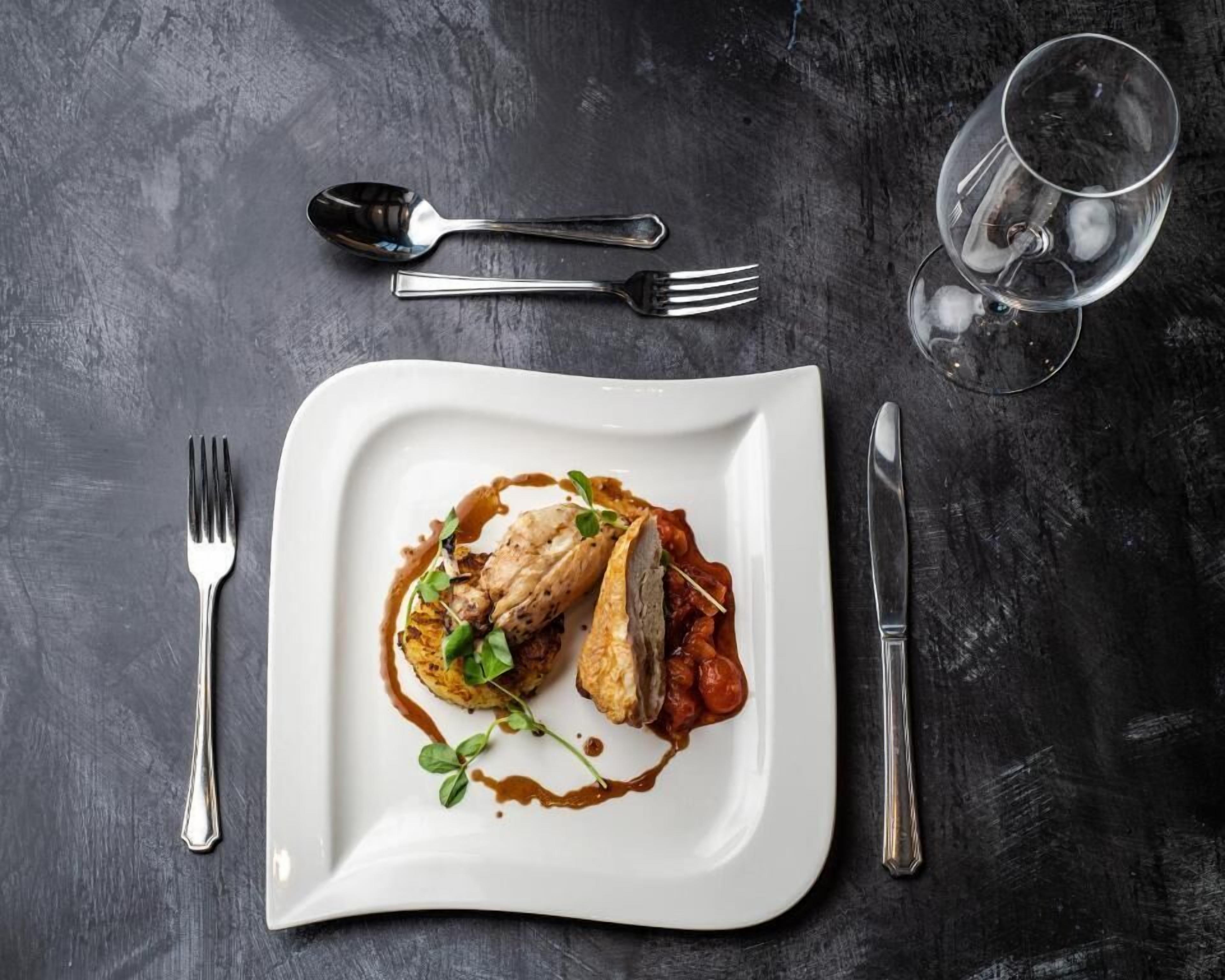 A white square plate with food, a fork, a knife, and a glass of water on a dark surface.