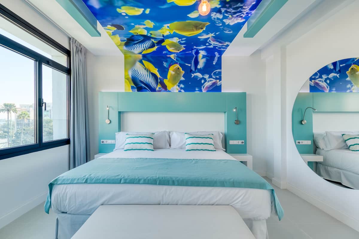 A modern hotel room with a large bed, a ceiling mural of tropical fish, and a view of palm trees.