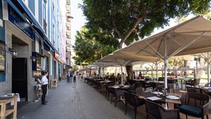 An outdoor café with tables and chairs under a canopy, a waiter standing by the entrance, and a building with a sign reading "Restaurante Santa Catalina" in the background. - HD Acuario Lifestyle (Las Palmas de Gran Canaria)