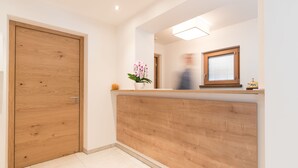 Zona de recepción con mostrador de madera, una orquídea en maceta y una ventana con persianas.