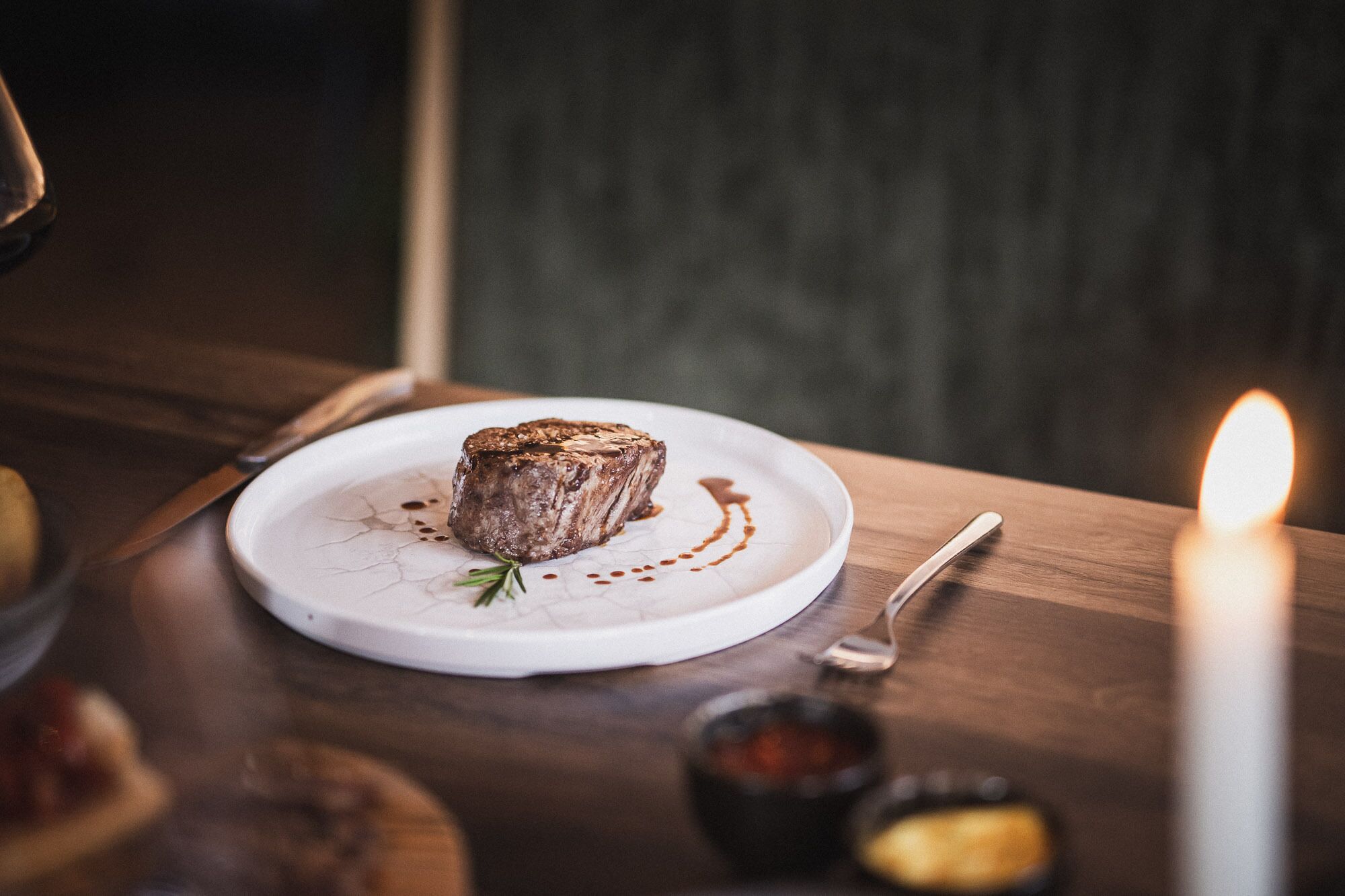 A steak on a plate with a candle and a spoon on a wooden table.