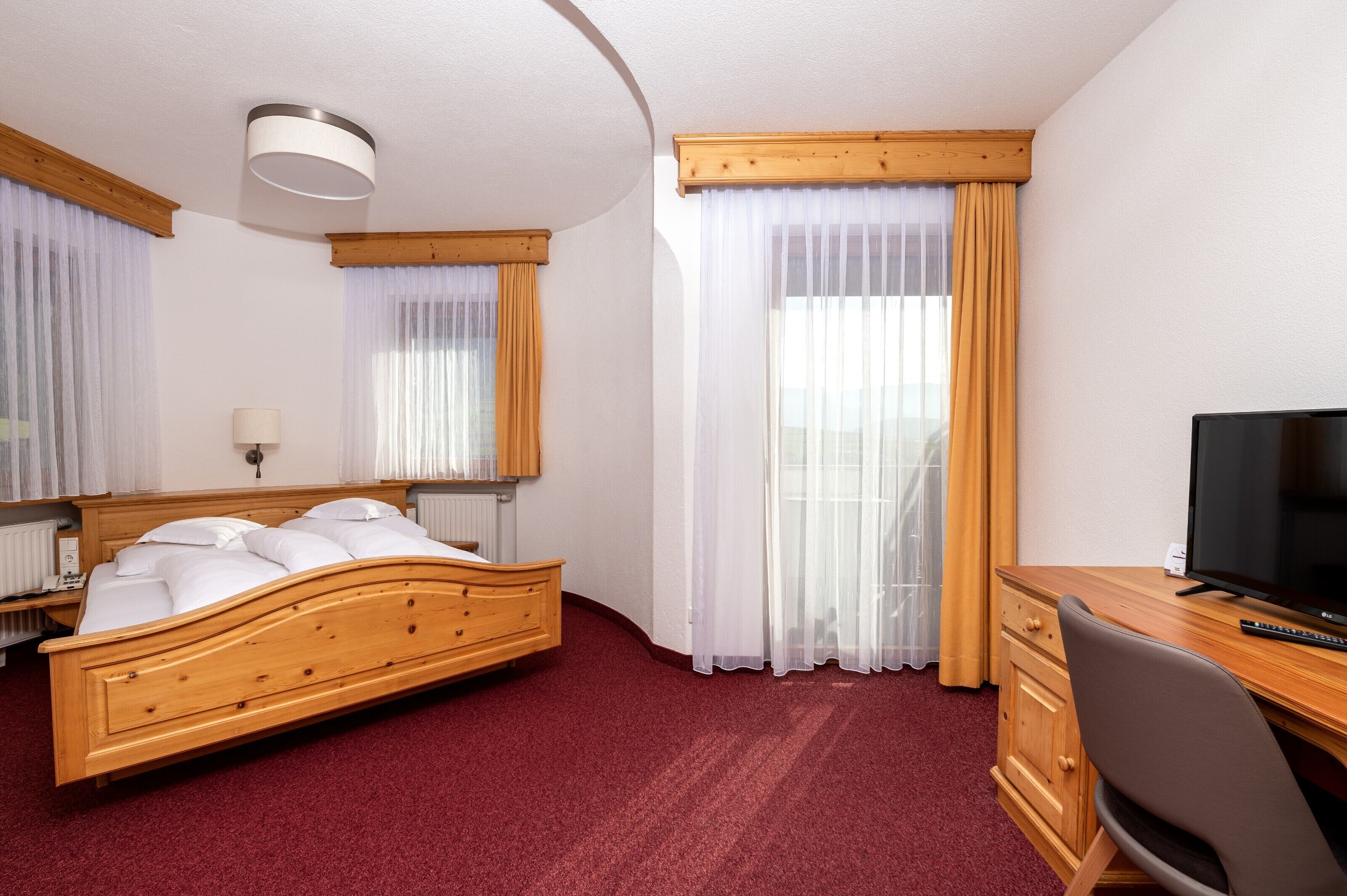 A hotel room with a wooden bed, a desk with a TV, and a window with sheer curtains.