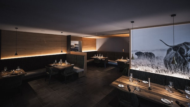 Un restaurante moderno con paredes de madera, suelo oscuro y una gran obra de arte iluminada de un perro. Las mesas están dispuestas con copas de vino y cubiertos.