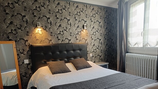 Una cama bien tendida con un cabecero oscuro y dos almohadas, una mesita de noche, un espejo y una ventana con cortinas.
