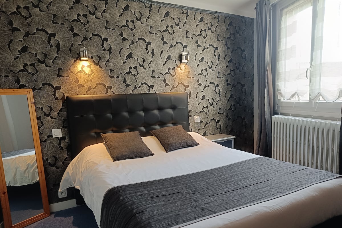 A neatly made bed with a dark headboard and two pillows, a bedside table, a mirror, and a window with curtains.