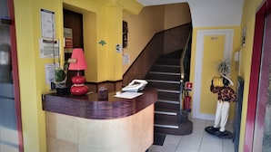 Zona de recepción de hotel con un mostrador curvo, un teléfono, una lámpara roja y una escalera que sube a otro piso.
