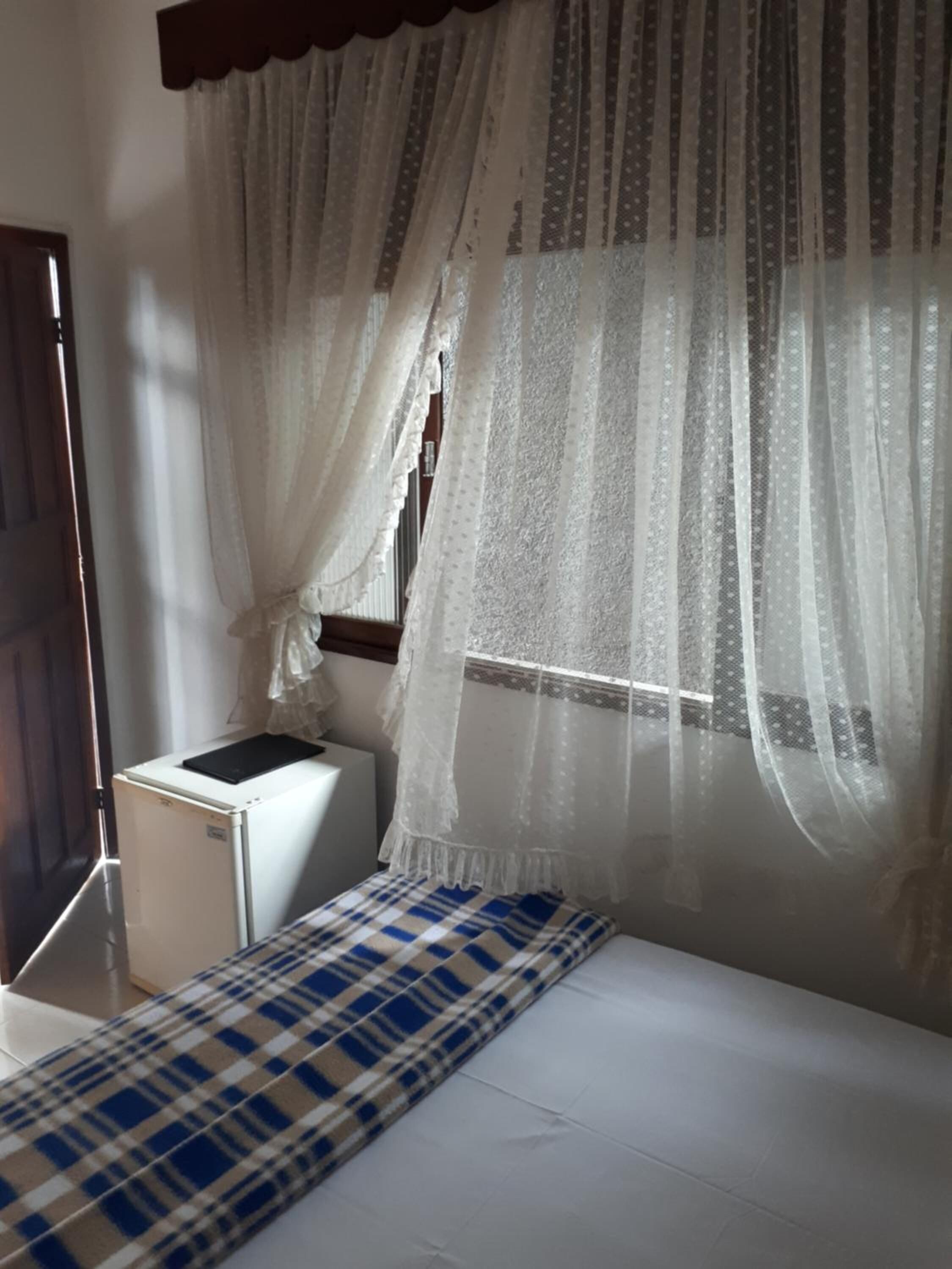 A bedroom with a bed, a small bedside table, a window with sheer curtains, and a door.
