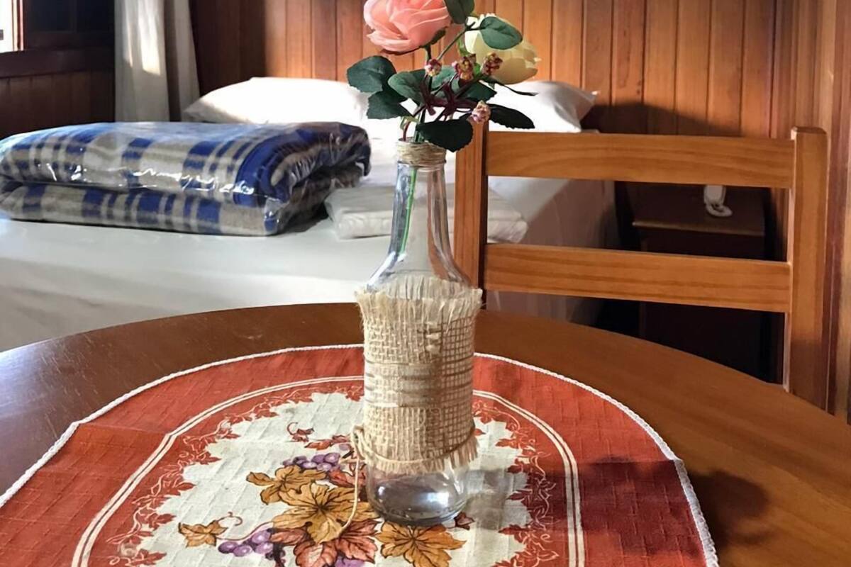 Ein Zimmer mit einer holzverkleideten Wand, einem Bett mit einer blau-weiß karierten Decke, einer Vase mit einer rosa Blume auf einem Tisch und einem Gemälde über dem Bett.