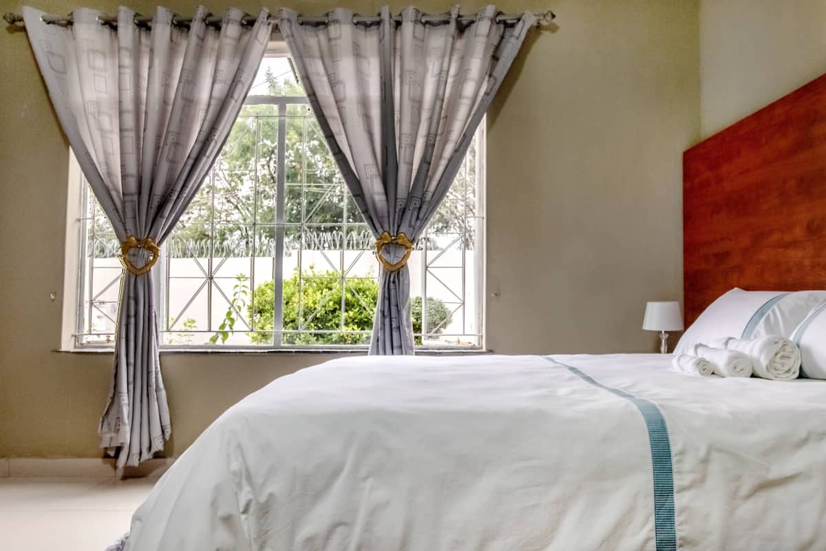 A hotel room with a large bed, a window with curtains, and a view of greenery outside.