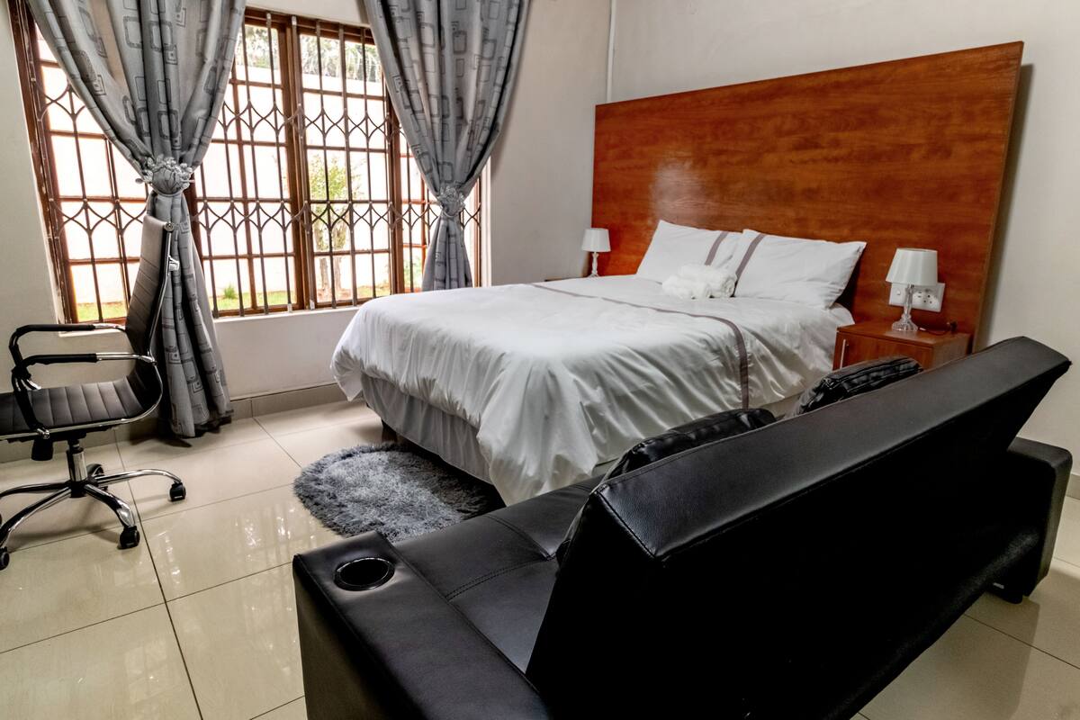 A bedroom with a large bed, a wooden headboard, two bedside tables with lamps, a black office chair, and a window with curtains.