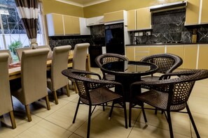 Un comedor con una mesa redonda y cuatro sillas de mimbre, una ventana con cortinas, y una cocina con armarios amarillos y un refrigerador negro.