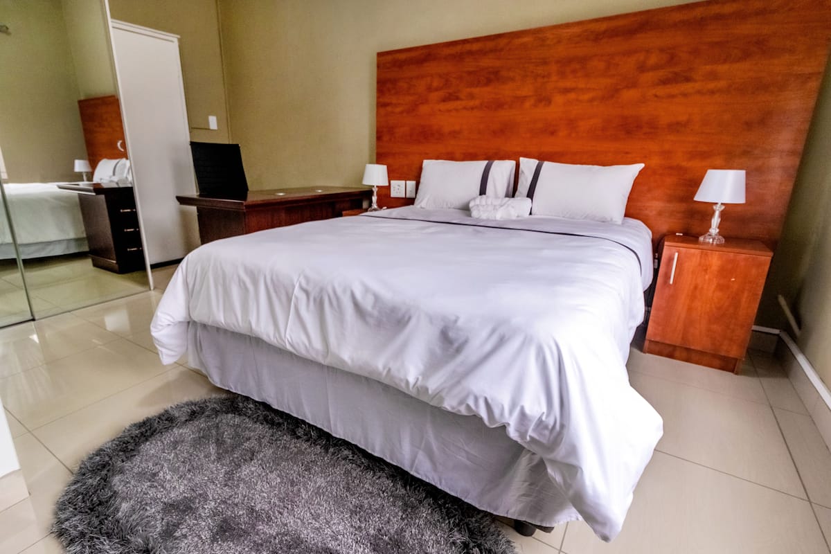A hotel room with a large bed, a wooden headboard, two bedside tables with lamps, a small desk with a chair, and a fluffy rug.