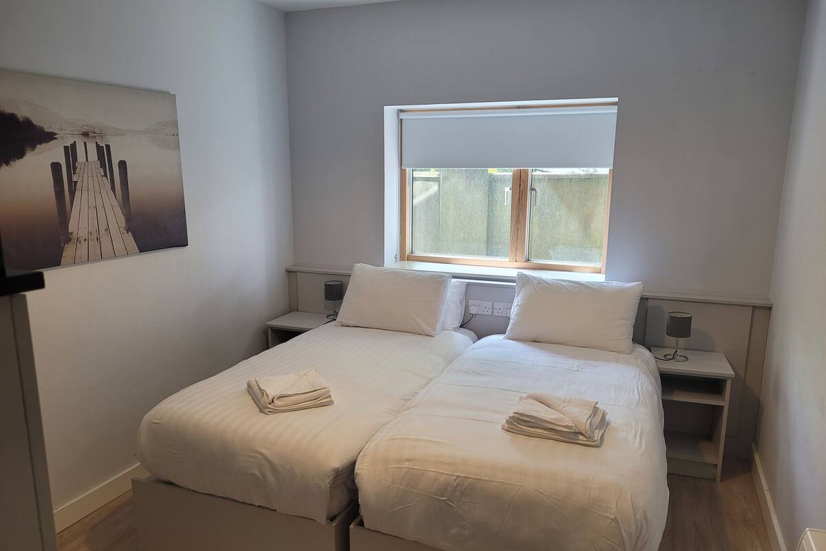 A double bed with two pillows and a folded towel, a bedside table with a lamp, a window with blinds, and a painting on the wall.