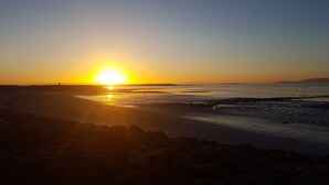 Una playa al atardecer, con el sol bajo en el horizonte, agua tranquila y primer plano rocoso.