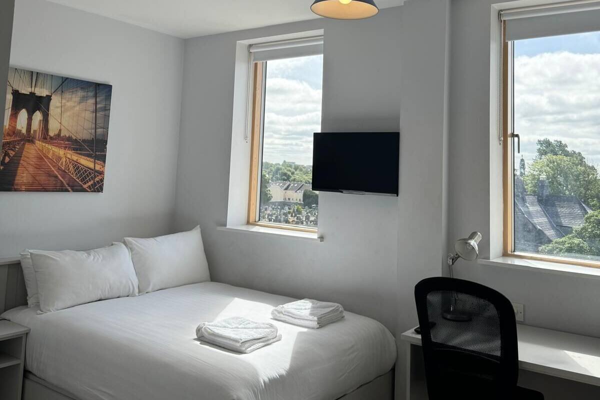 A bedroom with a bed, a desk, a chair, a TV, and windows with a view of a bridge and trees.