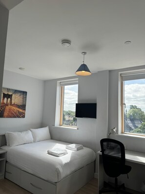 A bedroom with a bed, a desk, a chair, a TV, and windows with a view of a bridge and trees. - Swuite Galway (Galway)