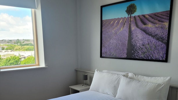 Una cama bien hecha con ropa blanca y toallas, una mesita de noche con lámpara, y un ventanal con vistas a un campo de lavanda y un árbol.