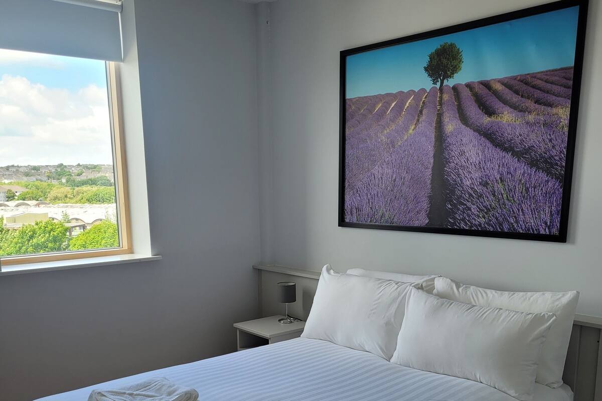 A neatly made bed with white linens and towels, a bedside table with a lamp, and a large window with a view of a lavender field and a tree.
