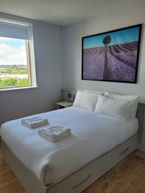 Una cama bien hecha con ropa blanca y toallas, una mesita de noche con lámpara, y un ventanal con vistas a un campo de lavanda y un árbol.