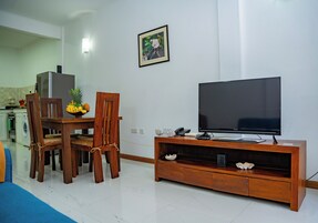 Una sala de estar con una mesa de comedor de madera, sillas, un televisor sobre un mueble de madera y una cocina al fondo.