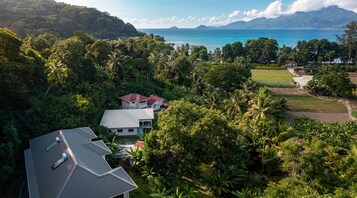 Una casa rodeada de exuberante vegetación tropical, con vista al océano y montañas al fondo.