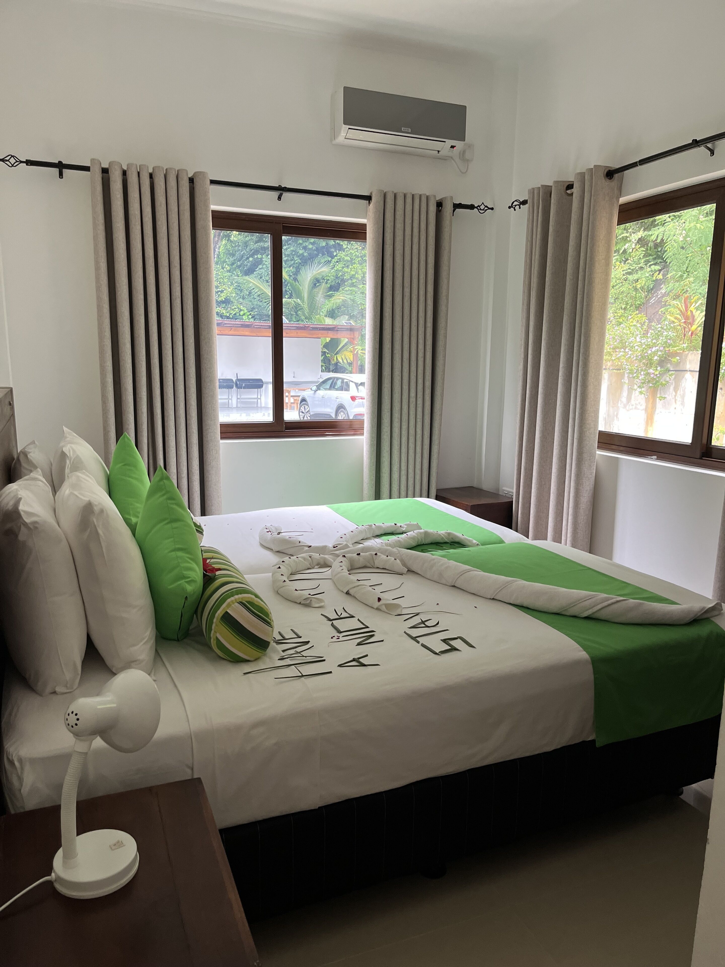 Ein Hotelzimmer mit Bett, Nachttisch mit Lampe, Fenster mit Vorhängen und Klimaanlage.