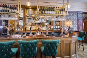 A bar area with multiple shelves of bottles and glasses, a bar counter, and three green upholstered bar stools. - Three Falcons Hotel (London)