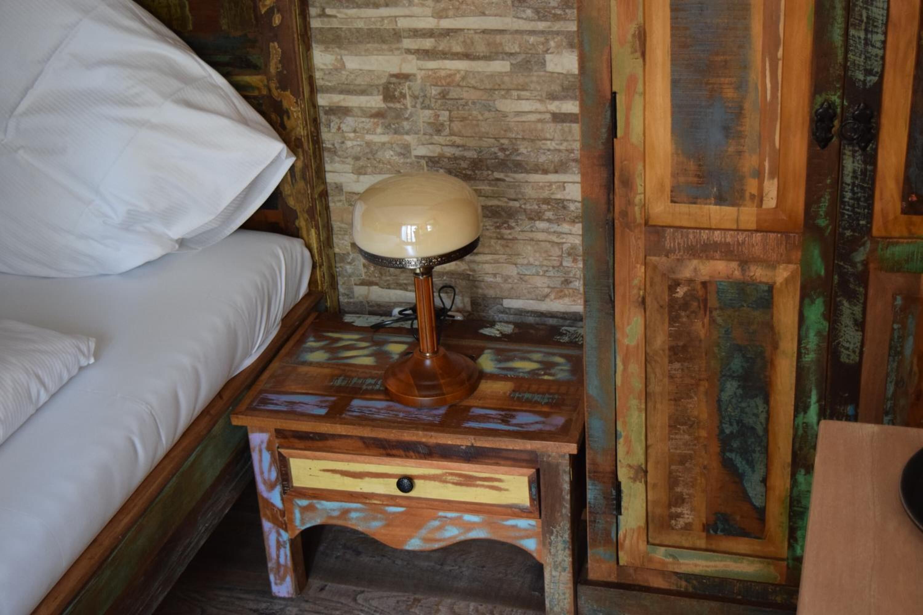 Una mesita de noche de madera con una lámpara, una cama con ropa blanca y una pared rústica con diseño de piedra.