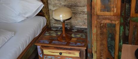 Una mesita de noche de madera con una lámpara, una cama con ropa blanca y una pared rústica con diseño de piedra.