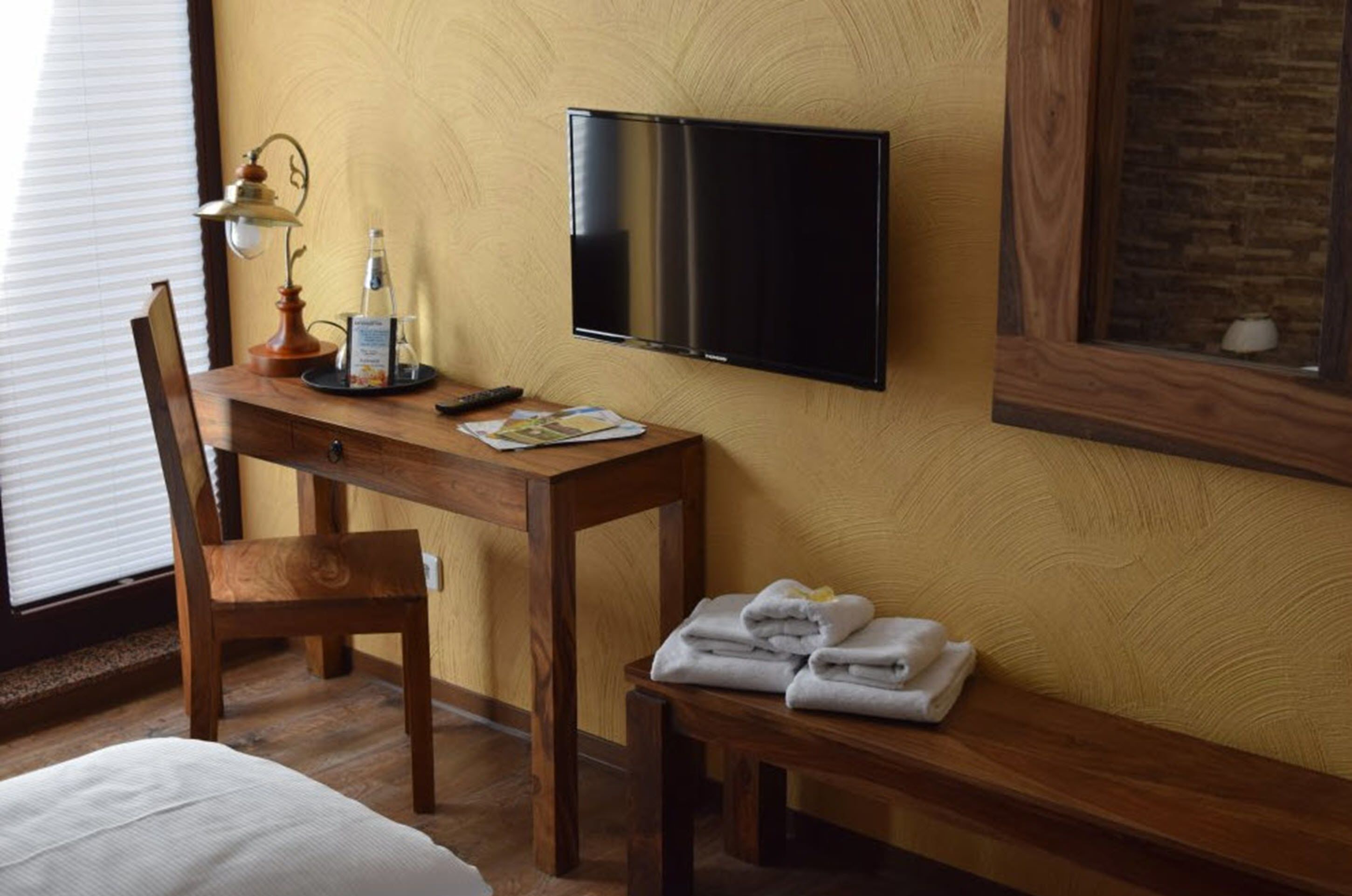 Habitación de hotel con un escritorio de madera, un televisor de pantalla plana montado en la pared, una silla, un banco con toallas y una lámpara.