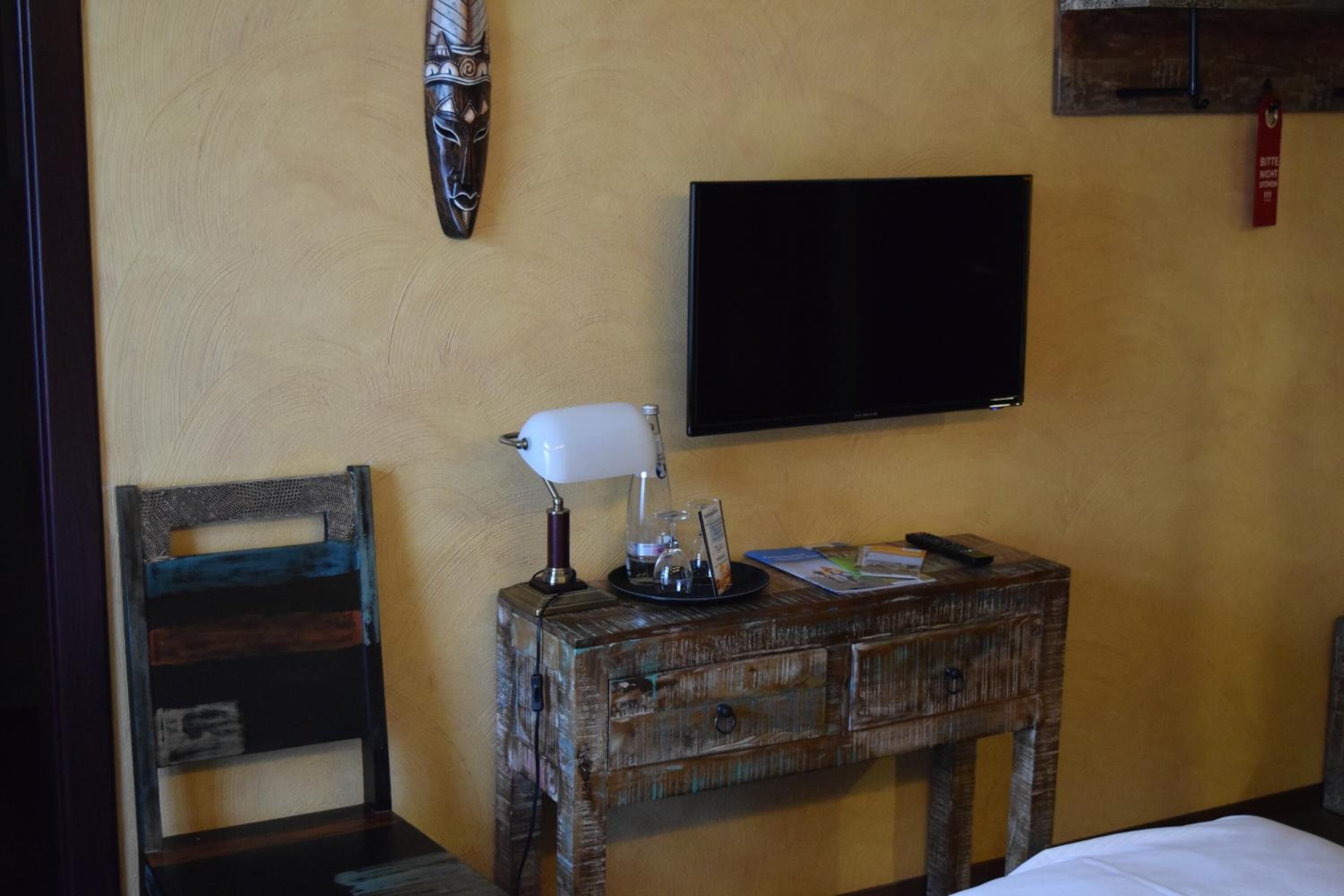 A room with a wooden nightstand, a white lamp, a flat-screen TV, and a chair.