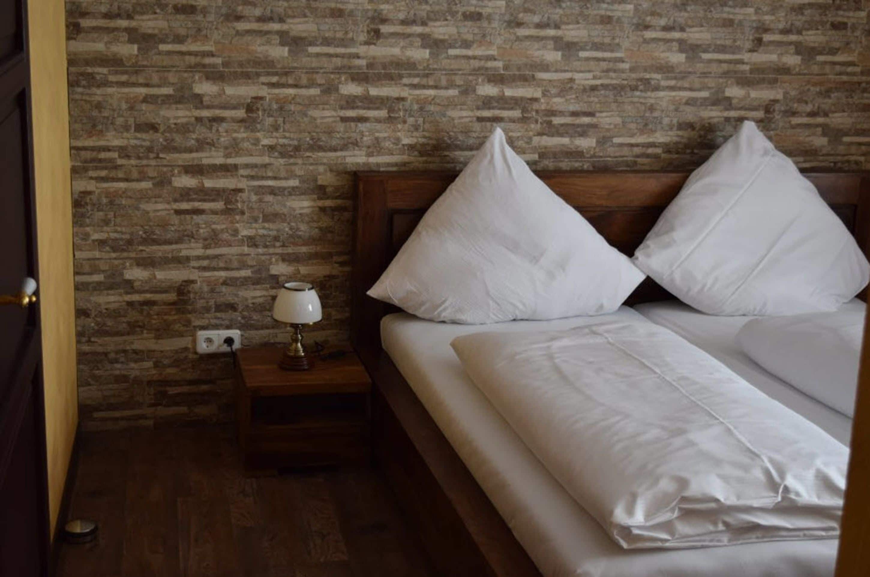 Une chambre avec un mur en pierre, une table de chevet en bois surmontée d’une lampe, et un lit avec une literie blanche.