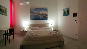 A hotel room with a bed, two bedside tables, a chair, a desk, and artwork on the walls. - Gymnasium (Syracuse)