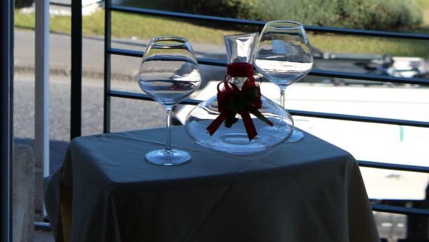 A table with two wine glasses and a decorative item, viewed through a window overlooking a parking area with cars and greenery. - Gymnasium (Syracuse)